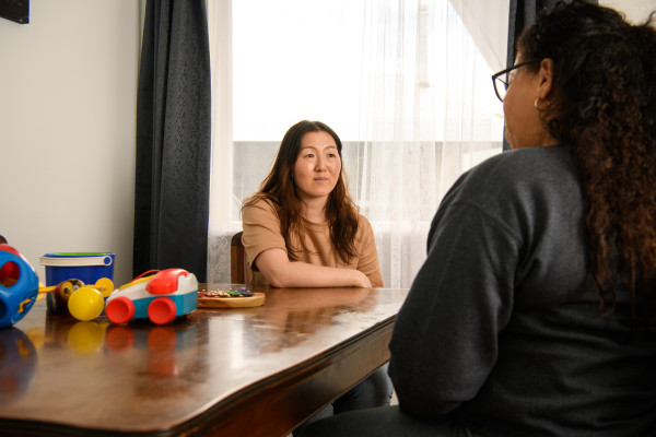 Two women talking at dining room table, with kids toys on top.